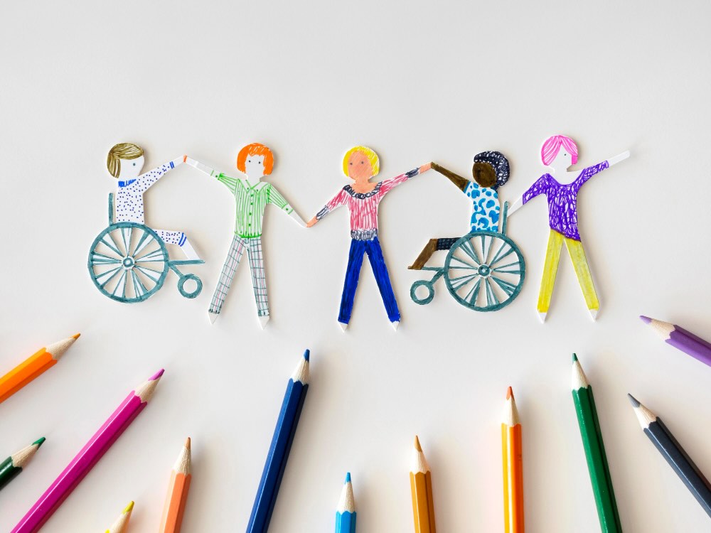 Inclusive community artwork celebrating International Day of People with Disability Paper cut-out figures, including people with and without disability, holding hands in a circle surrounded by coloured pencils, symbolising inclusion and community.