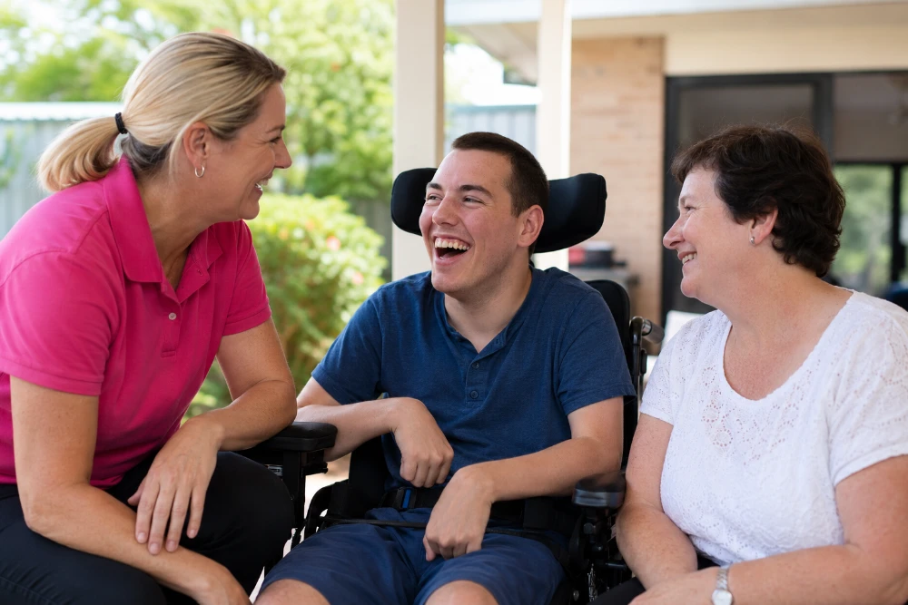 Support worker in pink polo shirt laughing with young man in wheelchair and older woman on outdoor patio, representing NDIS support and connection