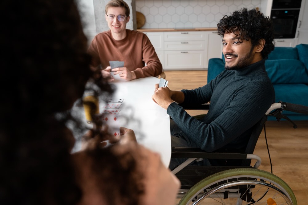 SDA home with SIL support showing independence and inclusion through NDIS housing depicted by a man using a wheelchair smiling while playing cards with friends